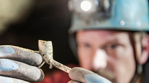 A caver wearing a blue helmet with a lit headtorch holds up a white clay pipe